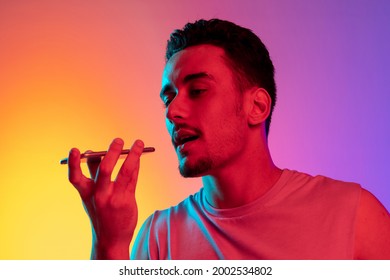 Recording Voice Message. Portrait Of Latina Young Man With Phone, Smartphone Isolated On Gradient Yellow Purple Background In Neon Light. Concept Of Emotions, Facial Expression.