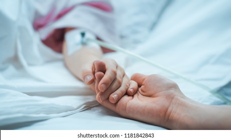 Recovering Little Child Lying In The Hospital Bed Sleeping, Mother Holds Her Hand Comforting. Focus On The Hands. Emotional Family Moment.