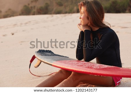 Recreation concept. Pensive relaxed young surfer focused aside, keeps hands together under chin, elbows on surfboard, admires beautiful sunset, poses in secluded place with quiet calm atmosphere