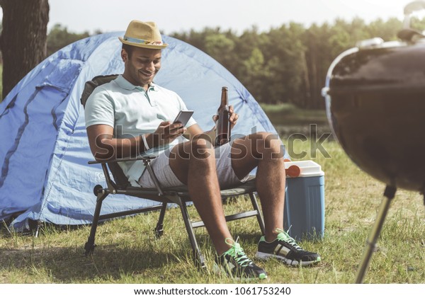 Recreation\
concept. Smiling guy with cellphone and bottle in hands sitting on\
folding chair. Camp tent on\
background