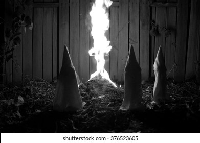 Recreation of a Klu Klux Klan meeting.  Hooded and robed, members stand around a burning cross at night.  Added film grain, documentary style.