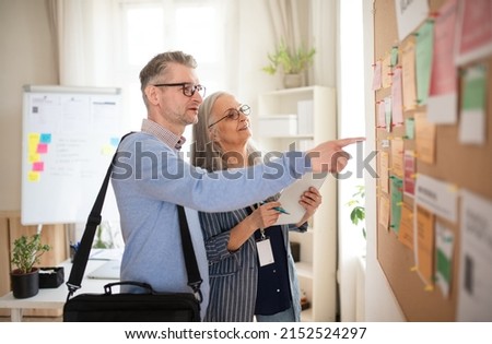 Recrutiment agency employee standing in front of employment noticeboard and helping mature man search for a job.