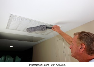 Rectangle White Intake Grid To A Furnace Being Dusted With A Microfiber Gray Dust Wand By An Adult Man. Mature Male Dusting A Rectangle Ceiling HVAC Intake Grid Of A Home Furnace To Reduce Allergens.