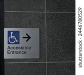 Rectangular accessible entrance sign, featuring blue wheelchair disability icon, black text, braille, and a black arrow on a dark wall