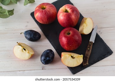 In a rectangular black dish on a white wooden table are three juicy beautiful ripe red apples of the early winter variety Gala. Next to the slices of cut fruit, a blue plum and a knife