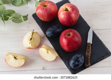 In a rectangular black dish on a white wooden table are three juicy beautiful ripe red apples of the early winter variety Gala. Next to the slices of cut fruit, a blue plum and  knife. Selective Focus