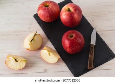 In a rectangular black dish on a white wooden table are three juicy beautiful ripe red apples of the early winter variety Gala. Next to the slices of the cut fruit and a knife. Top view
