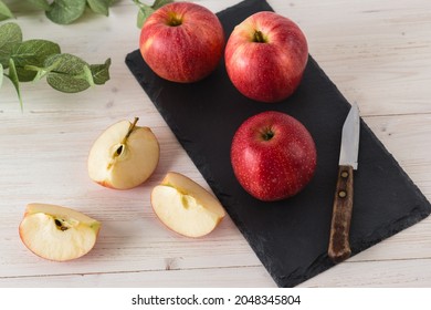 In a rectangular black slate dish on a white wooden table are three juicy beautiful ripe red apples of the early winter variety Gala. Next to the slices of the cut fruit and a knife. Top view