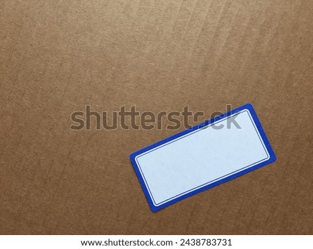 A rectangular blank white sticker with a blue frame is glued onto the ribbed surface of brown cardboard. Space for text