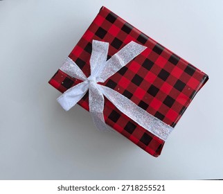 Rectangular Box Wrapped In Black And Red Patterned Christmas Paper And A White Glittery Ribbon Is Tied Around It Into A Bow Placed At An Angle On Top Of A White Smooth Background 