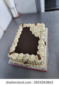 A rectangular cake featuring a smooth chocolate ganache center, decorative cream frosting borders, and chocolate bar pieces on a metal table.