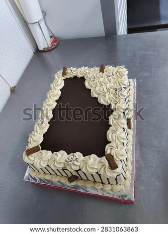 A rectangular cake featuring a smooth chocolate ganache center, decorative cream frosting borders, and chocolate bar pieces on a metal table.