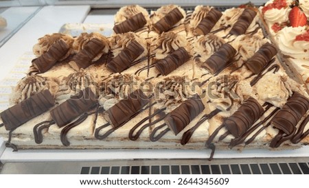 Rectangular cake with layers of cream, decorated with whipped cream rosettes, cocoa powder, chocolate drizzle, and sliced Kinder bars.