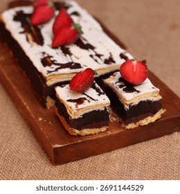 Rectangular chocolate brownie or cake with a thick white frosting layer, drizzled with chocolate sauce and topped with fresh strawberries, served on a rustic wooden board