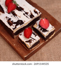Rectangular chocolate brownie or cake with a thick white frosting layer, drizzled with chocolate sauce and topped with fresh strawberries, served on a rustic wooden board