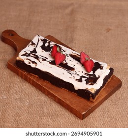 Rectangular chocolate brownie or cake with a thick white frosting layer, drizzled with chocolate sauce and topped with fresh strawberries, served on a rustic wooden board