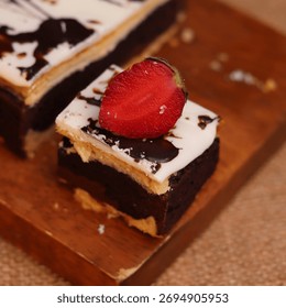 Rectangular chocolate brownie or cake with a thick white frosting layer, drizzled with chocolate sauce and topped with fresh strawberries, served on a rustic wooden board