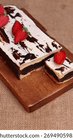 Rectangular chocolate brownie or cake with a thick white frosting layer, drizzled with chocolate sauce and topped with fresh strawberries, served on a rustic wooden board