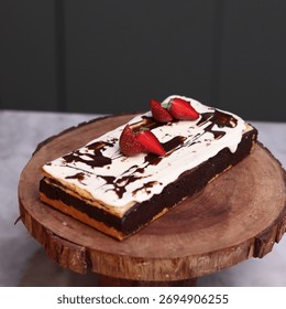 Rectangular chocolate brownie or cake with a thick white frosting layer, drizzled with chocolate sauce and topped with fresh strawberries, served on a rustic wooden board