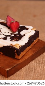 Rectangular chocolate brownie or cake with a thick white frosting layer, drizzled with chocolate sauce and topped with fresh strawberries, served on a rustic wooden board