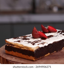 Rectangular chocolate brownie or cake with a thick white frosting layer, drizzled with chocolate sauce and topped with fresh strawberries, served on a rustic wooden board