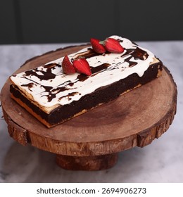 Rectangular chocolate brownie or cake with a thick white frosting layer, drizzled with chocolate sauce and topped with fresh strawberries, served on a rustic wooden board
