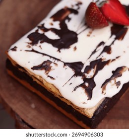 Rectangular chocolate brownie or cake with a thick white frosting layer, drizzled with chocolate sauce and topped with fresh strawberries, served on a rustic wooden board