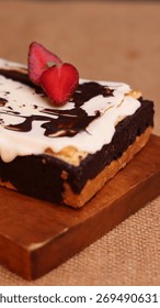 Rectangular chocolate brownie or cake with a thick white frosting layer, drizzled with chocolate sauce and topped with fresh strawberries, served on a rustic wooden board