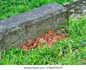 A rectangular concrete border sits nestled in vibrant green grass with a small pile of reddish-brown clay soil at its base. The close-up shot emphasizes the textured contrast between the aged gray masonry and the fresh earth in a garden setting.