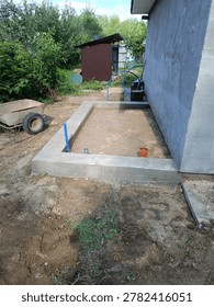 A rectangular concrete foundation sits adjacent to an existing grey building at a residential construction site. Inside the foundation walls, the ground is dirt, with orange and grey PVC plumbing pipes protruding upwards. A blue water hose lies nearby, and a metal wheelbarrow is parked on the dirt to the left. A small brown shed and green trees are visible in the background.