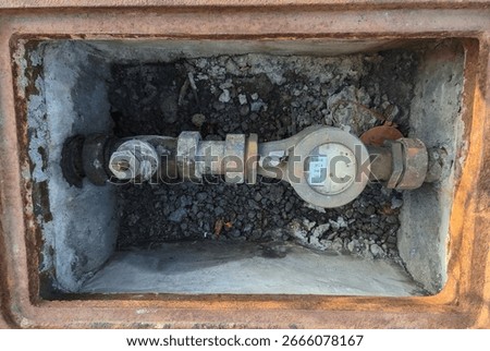 A rectangular concrete pit houses a water meter with visible mechanical parts, pipes, and fittings.