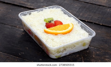 Rectangular container of fresh fruit salad topped with grated cheese and a slice of orange garnished with strawberries and kiwi