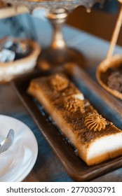 Rectangular dessert rests on a dark wooden tray. It features layered cake with piped cream rosettes. White plate holds a spoon beside it. Another chocolate dessert appears blurred in the background