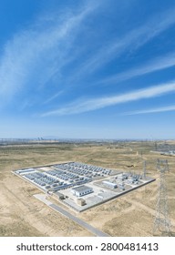 Rectangular fenced site with rows of gray battery containers, control buildings, and switchgear; surrounded by transmission towers, wind turbines, and open arid land under a bright blue sky. China