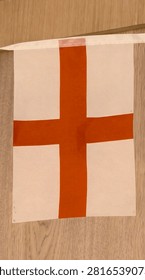 Rectangular flag featuring a bold red cross on a white background, symbolically associated with England. The flag hangs indoors against a wooden surface, emphasizing its cultural and historical significance. Perfect for themes of heritage, tradition, and national identity.