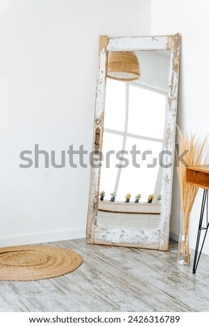 A rectangular fulllength mirror hangs on the wall of a room with hardwood flooring. The wood stain matches the door and window frames in the house