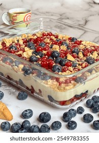 A rectangular glass dish filled with a layered dessert topped with berries and cookie crumbs. A cup of coffee and additional berries are scattered around.