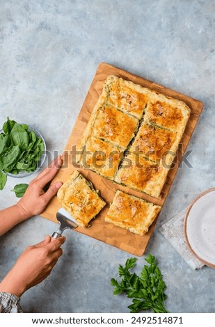 A rectangular Greek pie with spinach and cheese on a wooden board on a gray concrete background. Spinach recipes. Greek cuisine