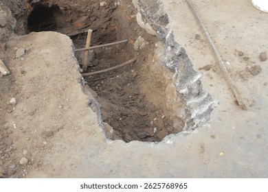 A rectangular hole dug into concrete and earth reveals layers of soil and a dark tunnel. Construction or repair work is evident, with exposed rebar and rough edges.