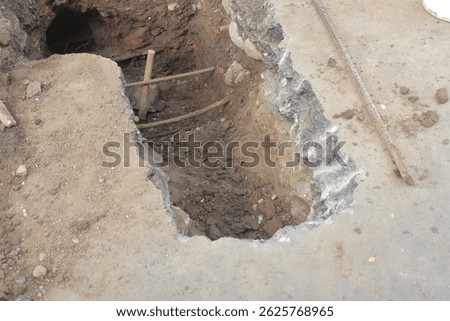 A rectangular hole dug into concrete and earth reveals layers of soil and a dark tunnel. Construction or repair work is evident, with exposed rebar and rough edges.