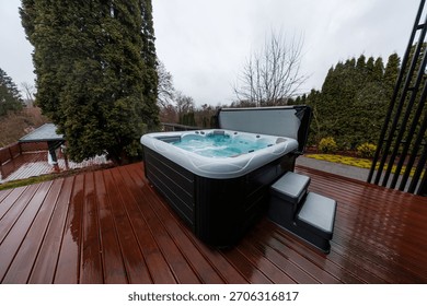 A rectangular hot tub churns on a wet timber deck, cover open and steps set. Steam rises in diffused light, evergreens and bare branches frame a cool overcast day.