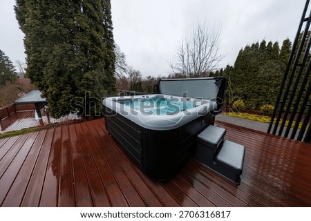 A rectangular hot tub churns on a wet timber deck, cover open and steps set. Steam rises in diffused light, evergreens and bare branches frame a cool overcast day.