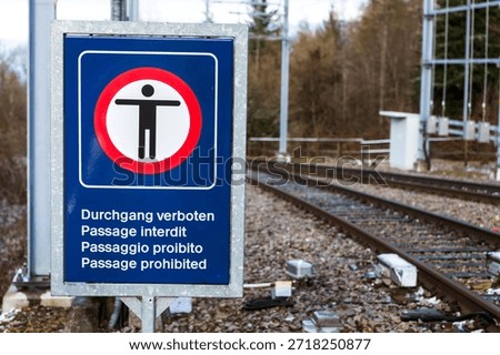 A rectangular metal sign with a dark blue background is mounted on a metal post beside railway tracks. The top section of the sign features a square red border enclosing a white circle with a black pictogram of a person with outstretched arms, indicating 