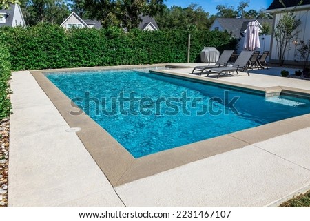 A rectangular new swimming pool with tan concrete edges in the fenced backyard of a new construction house with privacy hedges.