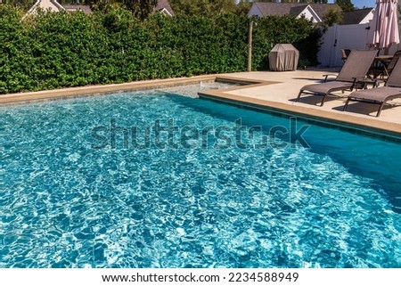 A rectangular new swimming pool with tan concrete edges in the fenced backyard of a new construction house with privacy hedges.