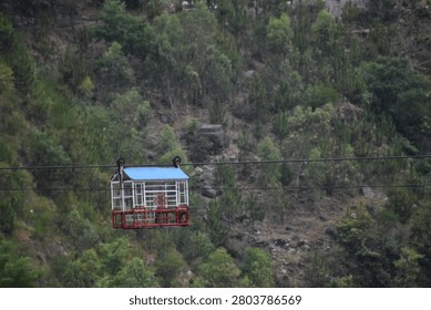 A rectangular, open-sided cable car cabin hangs suspended from a dark horizontal wire, glided high above an alpine valley floor. The aerial transport vehicle stands out against the wilderness with its distinct red lower frame, white structural bars enclosing the main compartment, and a light blue pitched roof.

The background is dominated by a steep, ascending mountain slope covered in a rugged mix of dry earth, grey stone outcroppings, and sparse green vegetation. Patches of wild shrubs and young deciduous trees line the craggy hillside, creating a highly textured and vertical backdrop.

Soft, uniform daylight filters through an overcast sky, casting an even, shadowless illumination over the entire scene. The immense scale of the steep mountain incline highlights the isolated and remote nature of the transit line crossing through the canyon wilderness.
