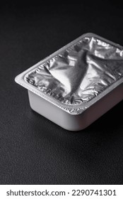 Rectangular plastic box with yogurt or cheese hermetically sealed with a foil lid on a dark concrete background