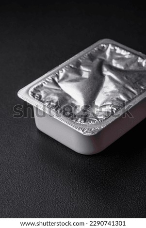 Rectangular plastic box with yogurt or cheese hermetically sealed with a foil lid on a dark concrete background