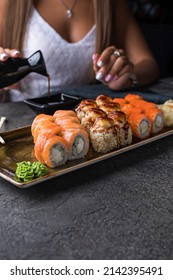 Rectangular plate with sushi rolls, wasabi and ginger, sushi sticks. In the background, a girl in white pours soy sauce into a saucepan. Close up.