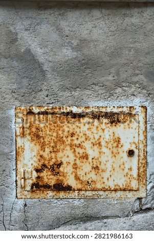 A rectangular rusty metal utility door with hinges and a lock is installed in a textured gray concrete wall surface.